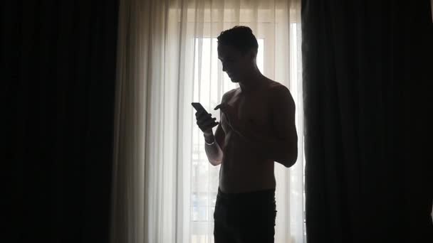 Jongeman texting op smartphone door raam thuis — Stockvideo