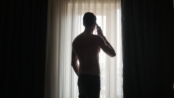 Young man talking on cell phone by window at home — Stock Video