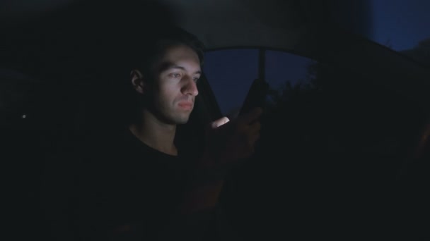Νεαρός άνδρας χρησιμοποιώντας ένα έξυπνο τηλέφωνο, ενώ κάθεται σε ένα αυτοκίνητο στο δρόμο τη νύχτα πόλη — Αρχείο Βίντεο