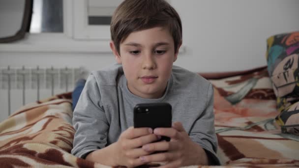 Niño jugando con el teléfono móvil en casa. Niño usando Smartphone acostado en una cama — Vídeo de stock