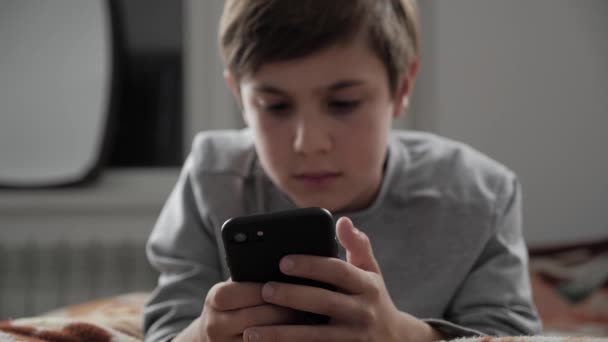 Niño jugando con el teléfono móvil en casa. Niño usando Smartphone mientras está acostado en una cama — Vídeos de Stock