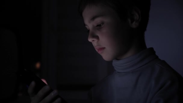 Ein fröhlicher Junge spielt begeistert auf einem Smartphone in einem Spiel im Dunkeln zu Hause. das Gesicht des Kindes wird von einem hellen Monitor beleuchtet — Stockvideo