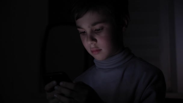 Happy Boy juega con entusiasmo en un teléfono inteligente en un juego en la oscuridad en casa. La cara del niño está iluminada por un monitor brillante — Vídeos de Stock