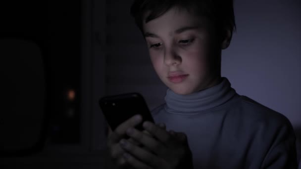 Ein fröhlicher Junge spielt begeistert auf einem Smartphone in einem Spiel im Dunkeln zu Hause. das Gesicht des Kindes wird von einem hellen Monitor beleuchtet — Stockvideo