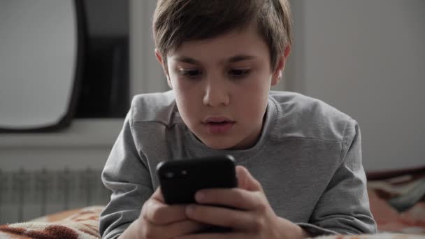 Παιδί αγόρι παίζει με το κινητό τηλέφωνο στο σπίτι. Παιδί χρησιμοποιώντας Smartphone ενώ ξαπλωμένος σε ένα κρεβάτι — Αρχείο Βίντεο