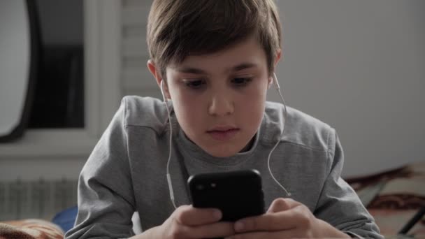 Детский мальчик играет с мобильным телефоном с наушниками дома. Ребенок с помощью смартфона, лежащий на кровати — стоковое видео