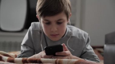 Çocuk çocuk cep telefonu kulaklık ile birlikte evde oynamak. Çocuk bir yatakta yatarken Smartphone kullanarak