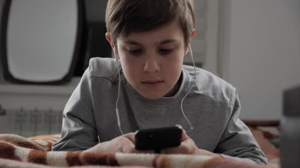 Niño jugando con el teléfono móvil con auriculares en casa. Niño usando Smartphone mientras está acostado en una cama — Vídeo de stock