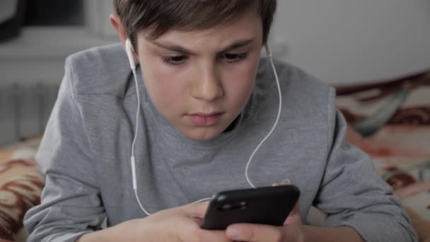 Niño jugando con el teléfono móvil con auriculares en casa. Niño usando Smartphone mientras está acostado en una cama — Vídeo de stock