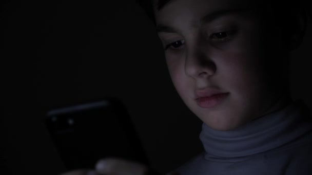 Happy Boy juega con entusiasmo en un teléfono inteligente en un juego en la oscuridad en casa. La cara del niño está iluminada por un monitor brillante — Vídeos de Stock