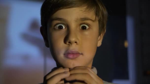 Kleiner verängstigter Junge, der im dunklen Raum Horrorfilme oder Videos auf dem Fernseher oder Computer anschaut. Spiegelung auf seinem Gesicht. — Stockvideo