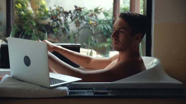 Junger Mann liegt im Badezimmer und arbeitet mit Laptop — Stockvideo