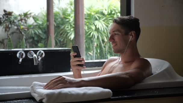 Jonge, gelukkige man liggend in badkuip luisteren naar muziek op smartphone in badkamer — Stockvideo