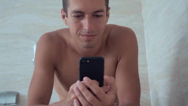 Mladý muž používá chytrý telefon sedí na WC doma. Muž sedí na WC s telefonem. — Stock video