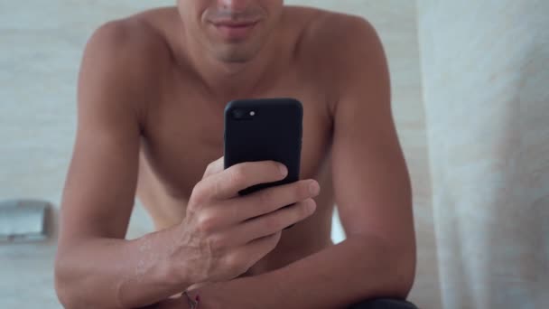 Le jeune homme utilise un téléphone intelligent assis sur les toilettes à la maison. Homme assis dans les toilettes avec téléphone . — Video