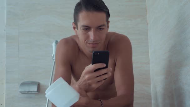 Молодий чоловік використовує смартфон, сидячи в туалеті вдома. Чоловік сидить у туалеті з телефоном . — стокове відео
