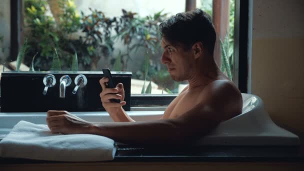 Dühös ember használ Smartphone fekve, a fürdőszobában fürdőkád. Bosszús ember dob egy okostelefon a vízbe — Stock videók