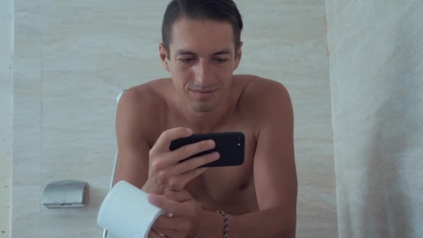 Le jeune homme utilise un téléphone intelligent assis sur les toilettes à la maison. Homme assis dans les toilettes avec téléphone . — Video