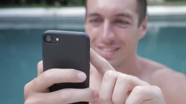 Attractive and handsome young man looks, scrolls through social media feed on smartphone while resting at pool on vacation — Stock Video