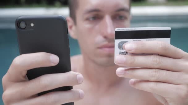 Joven comprando bienes de Internet en su teléfono inteligente con su tarjeta de crédito mientras nada en la piscina de vacaciones — Vídeo de stock