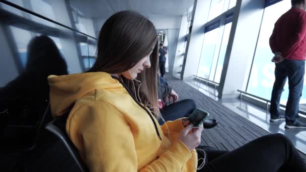 Жінка чекає на посадку в аеропорту, проводячи час, дивлячись фільм на смартфон. Дівчина з навушниками дивиться на екран мобільного пристрою — стокове відео
