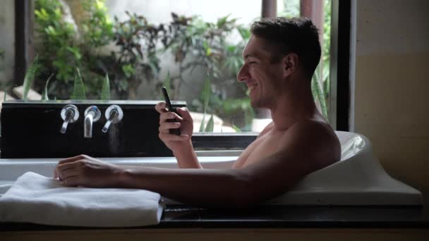 Улыбающийся молодой человек с помощью смартфона во время лежания в ванной комнате — стоковое видео