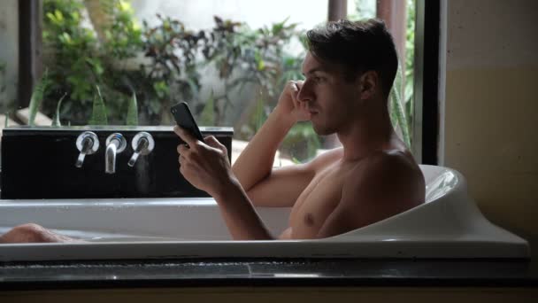 年轻人把智能手机掉进浴缸里的水里 马虎男子在浴室的水中丢失了一部智能手机 — 图库视频影像