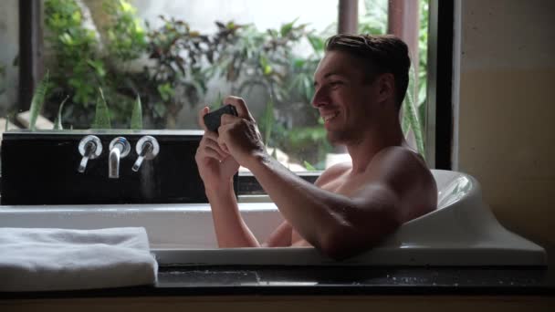 Joven hombre viendo vídeo en el teléfono inteligente mientras está acostado en la bañera en el baño — Vídeos de Stock