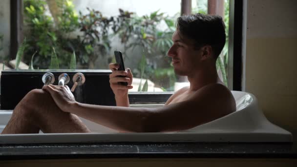 Mosolygó fiatal ember használ Smartphone fekve, a fürdőszobában fürdőkád — Stock videók