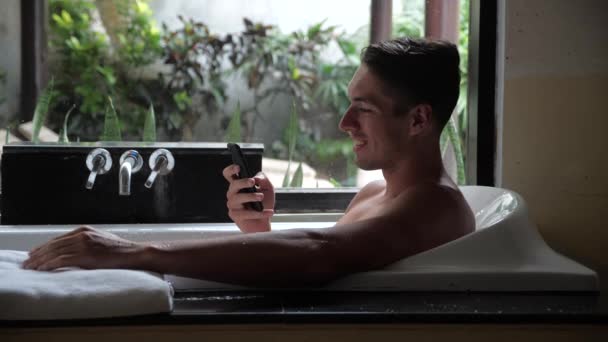 Sorridente giovane utilizzando Smartphone mentre si trova nella vasca da bagno in bagno — Video Stock