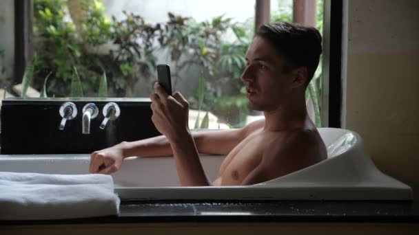 Mosolygó fiatal ember használ Smartphone fekve, a fürdőszobában fürdőkád. Az ember egyre jó üzenet egy okostelefon. Öröm és a gyönyör — Stock videók