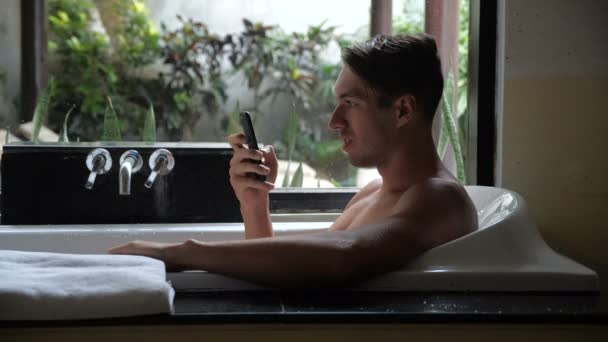 Giovane bell'uomo sdraiato nella vasca da bagno, usando lo smartphone, il successo nell'affare, sorridendo. Uomo ottenere messaggio win su smartphone mentre si trova in bagno — Video Stock