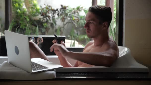 Drukke multitasking zakenman ligt in de badkamer en werkt met een laptop en een smartphone — Stockvideo