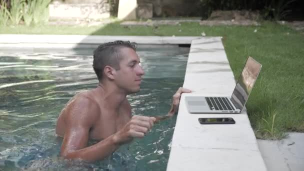 Jonge man aan het werk op laptop zitting in het zwembad. De kerel freelancer werkt in een prachtige plek in het resort bij het zwembad — Stockvideo