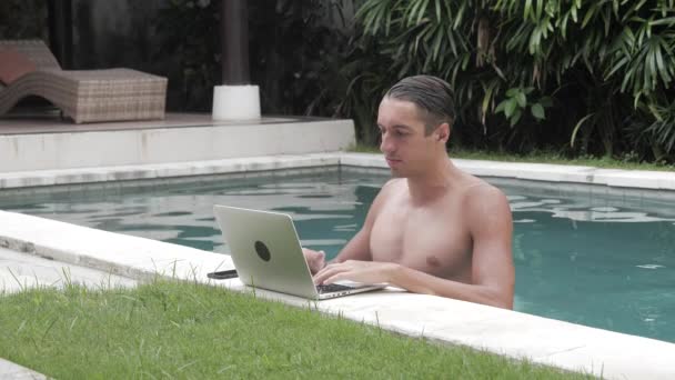 Jonge man aan het werk op laptop zitting in het zwembad. De kerel freelancer werkt in een prachtige plek in het resort bij het zwembad — Stockvideo