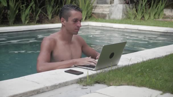 Mladý muž pracuje na notebooku v bazénu. Chlap na volné noze pracuje na krásném místě v areálu u bazénu — Stock video
