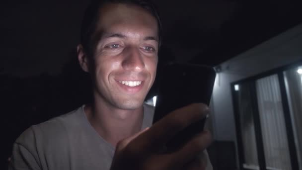 Joven hombre sonriente utiliza un teléfono inteligente por la noche en frente de la casa . — Vídeos de Stock