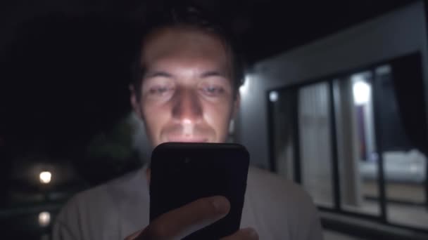 Jovem sorridente homem usa um smartphone à noite na frente da casa . — Vídeo de Stock