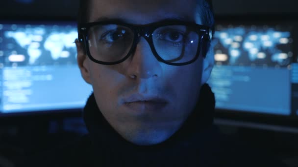 Portrait of Hacker programmer in glasses looks at camera while blue code characters reflect on his face in cyber security center filled with display screens. — Stock Video