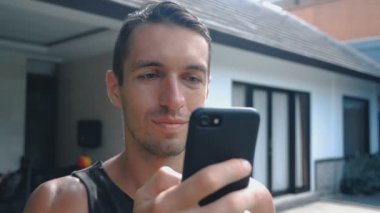 Portre, gülümseyen genç adam kullanarak Internet veya sosyal ağlar kontrol avlu ya da bahçe arka plan tarama Smartphone,
