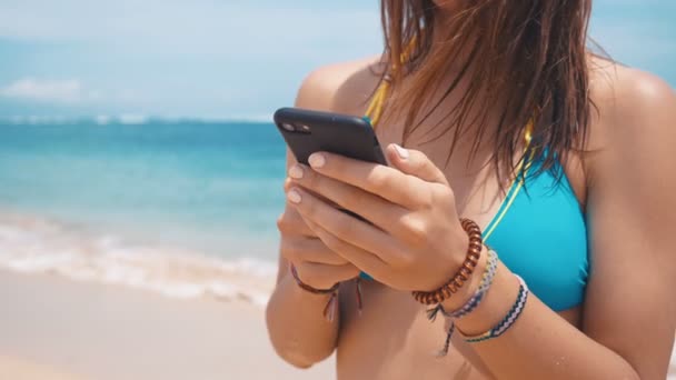 若い女性がビーチで海を背景にスマート フォンを使用しています。笑顔の女性観光客は、バリでの休暇中に水着. — ストック動画