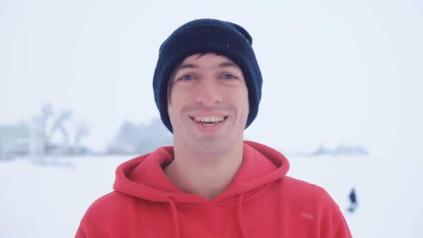 Портрет молодого человека в черной кепке смотрит в камеру, улыбаясь зимой снежным холодом на открытом воздухе — стоковое видео
