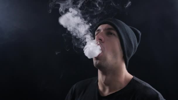 ชายหนุ่ม vaper หายใจเมฆใหญ่ของควันกับบุหรี่อิเล็กทรอนิกส์ vape บนพื้นหลังสีดําในการเคลื่อนไหวช้า — วีดีโอสต็อก