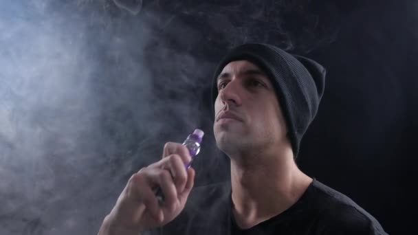 Young man vaper exhaling big clouds of smoke with e-cigarette vape on black background in slow motion — Stock Video