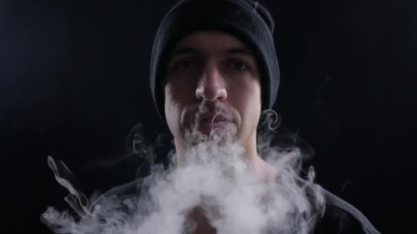 Молодой человек vaper выдыхая большие облака дыма с электронной сигаретой vape на черном фоне в замедленной съемке — стоковое видео