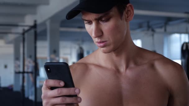 Atleta comprueba Smartphone mientras hace ejercicio. Hombre joven escribiendo descanso durante el entrenamiento en el gimnasio — Vídeo de stock