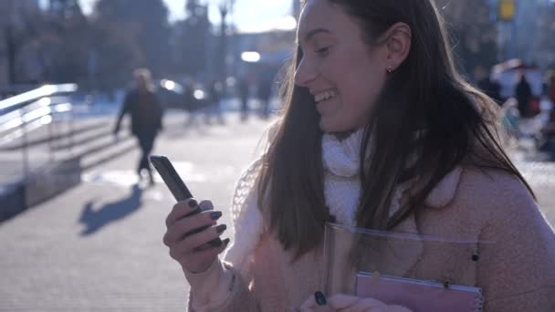 세련 된 코트와 스카프, 도시 거리에 웃는 갈색 머리 여자의 초상화 unsing 스마트폰 — 비디오