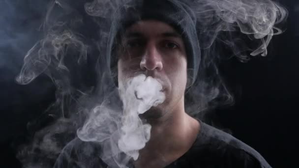 Jovem vaper exalando grandes nuvens de fumaça com vape e-cigarro no fundo preto em câmera lenta — Vídeo de Stock
