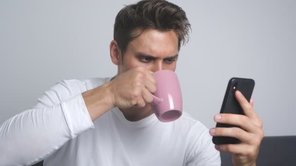 Homem sorridente atraente usando smartphone enviando mensagem nas mídias sociais e bebendo café em casa — Vídeo de Stock