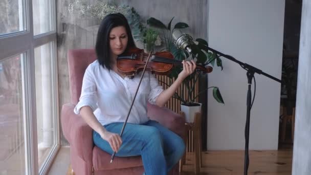 En violinist sitter på en stol spelar en fiol nära ett fönster i ett rum, stadig cam Shot, slow motion. — Stockvideo